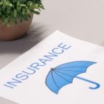 Image of paper with the word insurance and an umbrella printed on it, next to a laptop with someone's hand typing